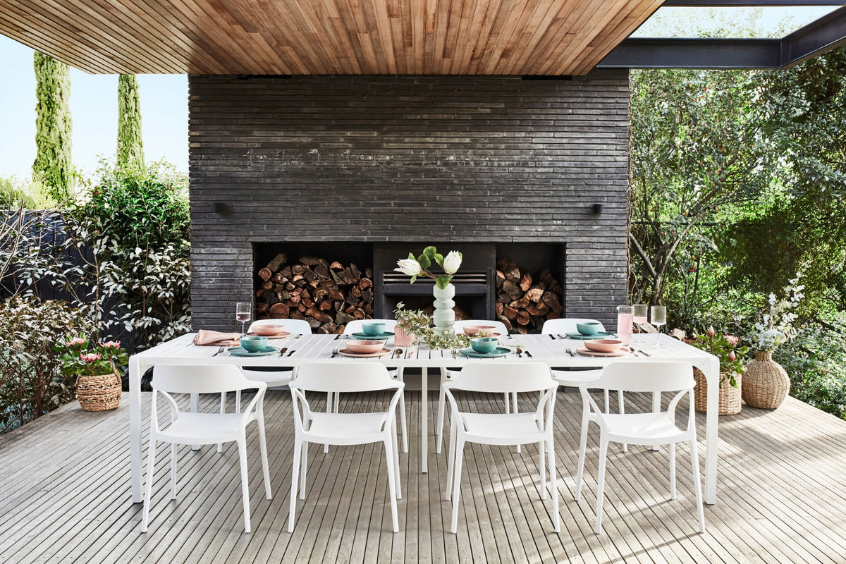 An outdoor patio features the Kace Dining Table 300cm White with 8 Inigo Outdoor Dining Chair White Package, set with green and pink tableware. A black brick wall with firewood storage highlights the background, surrounded by lush greenery.
