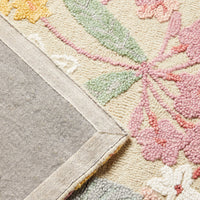 A corner of the Botanic Native Rug 240x150cm showcases its beige-bordered floral design in pink, green, and yellow tones. The hand-tufted rug, made from 100% wool, is partially flipped to show the gray underside.