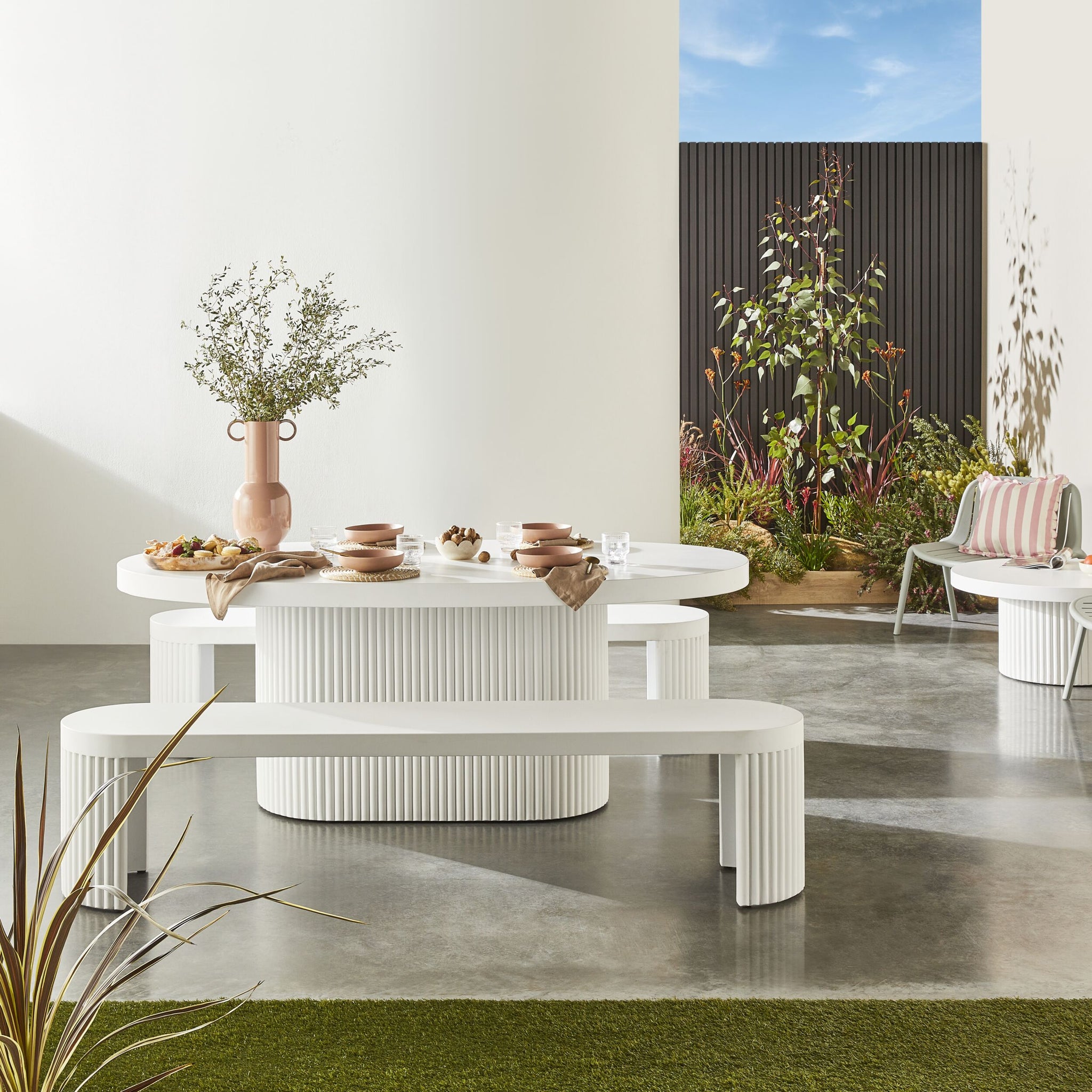 Transform your concrete patio into a stylish haven with the modern elegance of our Ripple Curved Outdoor Dining Table 200cm and Bench Package, featuring sleek white ribbed furniture, lush greenery, and contemporary tableware.