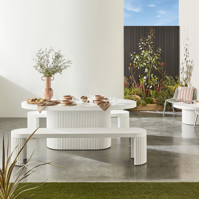 Transform your concrete patio into a stylish haven with the modern elegance of our Ripple Curved Outdoor Dining Table 200cm and Bench Package, featuring sleek white ribbed furniture, lush greenery, and contemporary tableware.