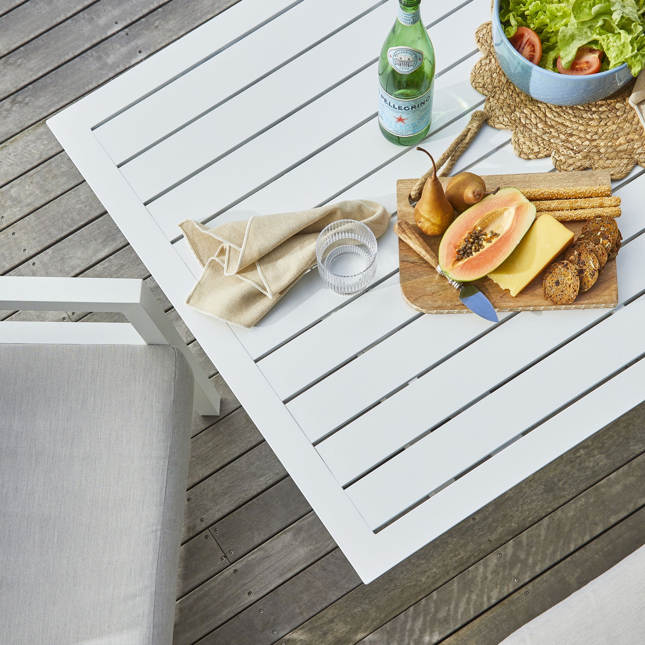 Close-up of a white outdoor dining table with a cheese board, fruit, bread, and a napkin, showing table slats and natural tones.