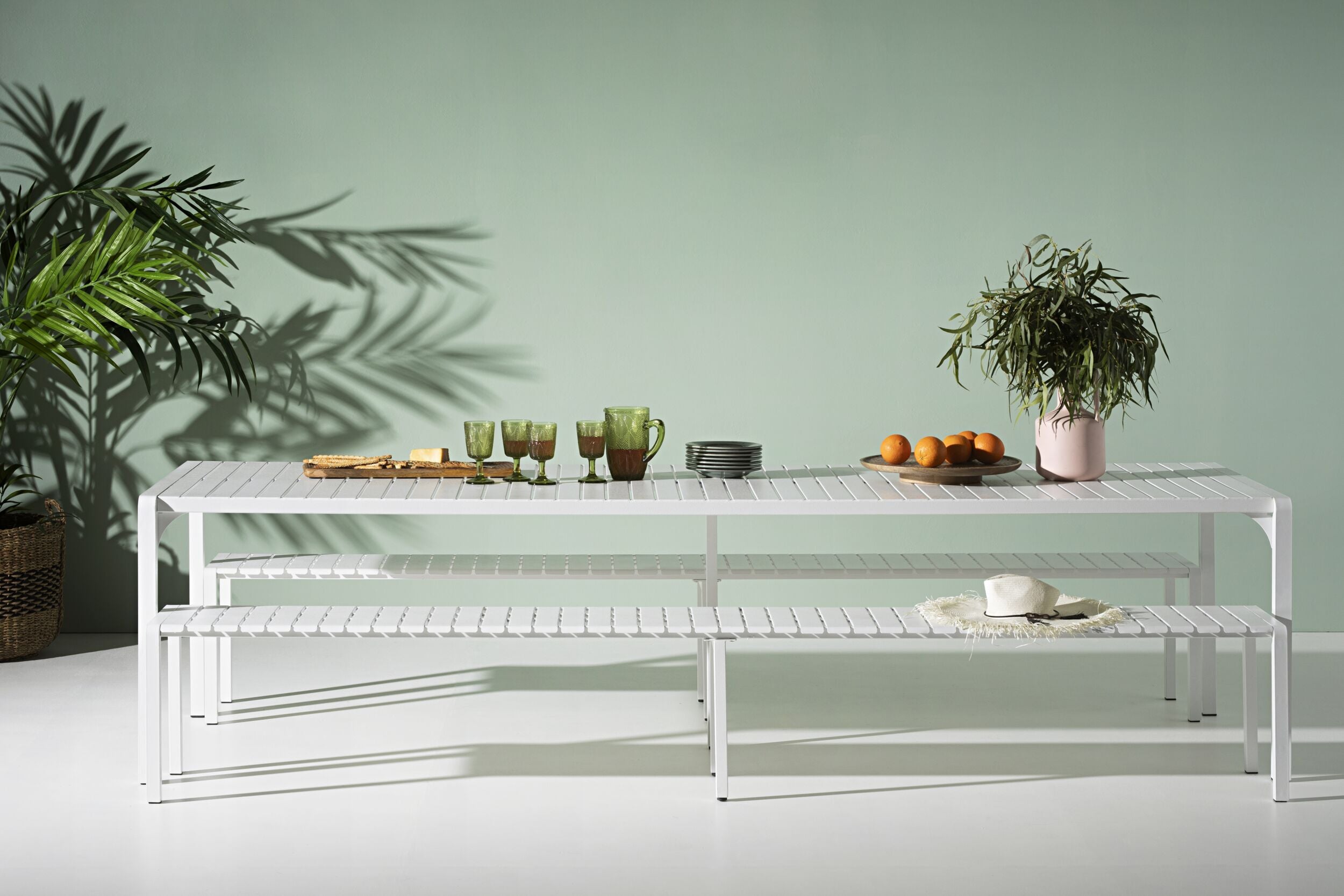 The Early Settler Kace Dining Table 300cm White with 2 Kace Dining Bench 284cm White Package showcases a white metal table and benches, elegantly decorated with green glassware, stacked plates, a potted plant, and oranges against a light green wall. Charming patterns are cast by shadows from a palm plant, making it an ideal choice for any alfresco entertaining area.