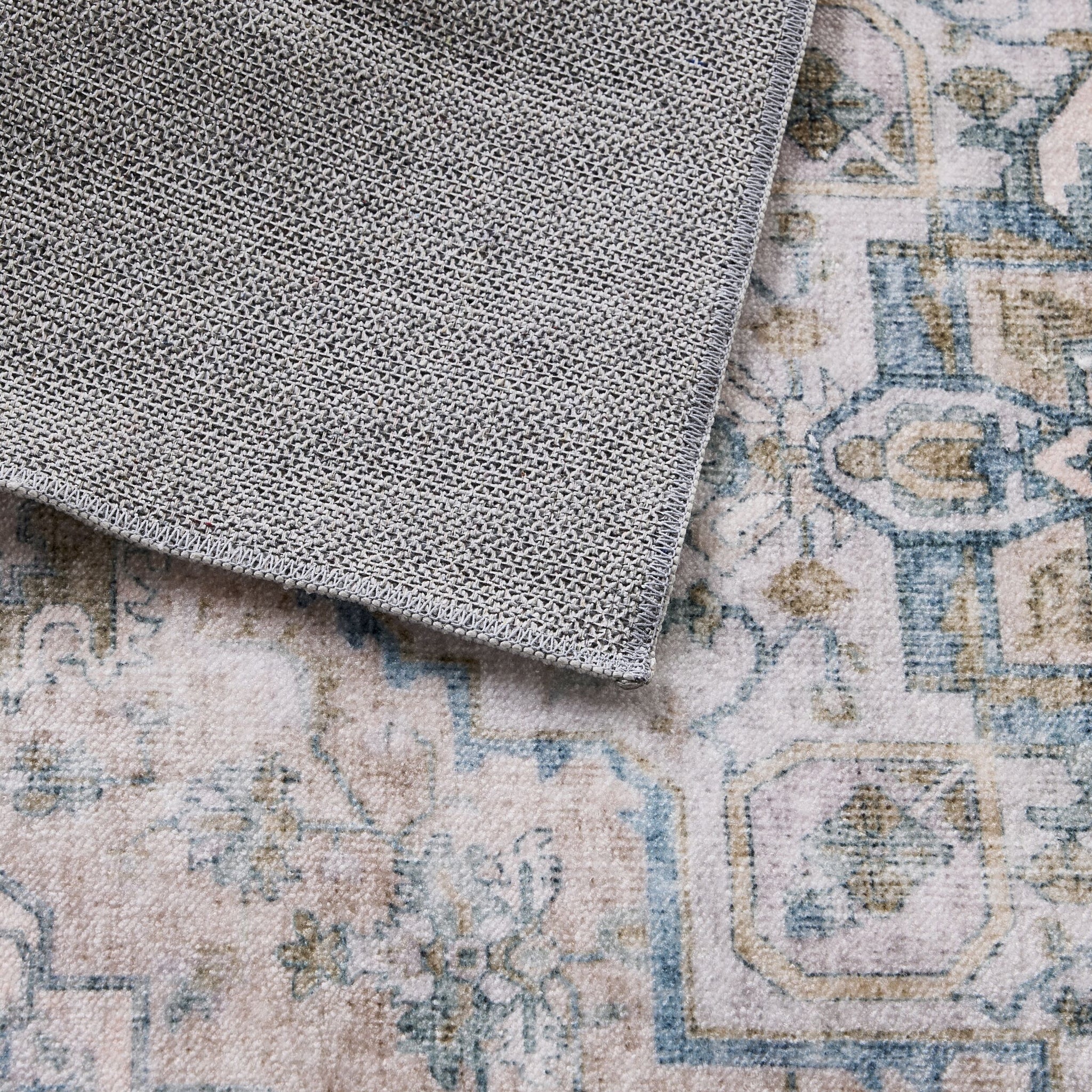 A close-up of the Manisa Washable Runner 80x200cm in gray textured fabric overlapping a patterned carpet with muted blue, beige, and gold tones.