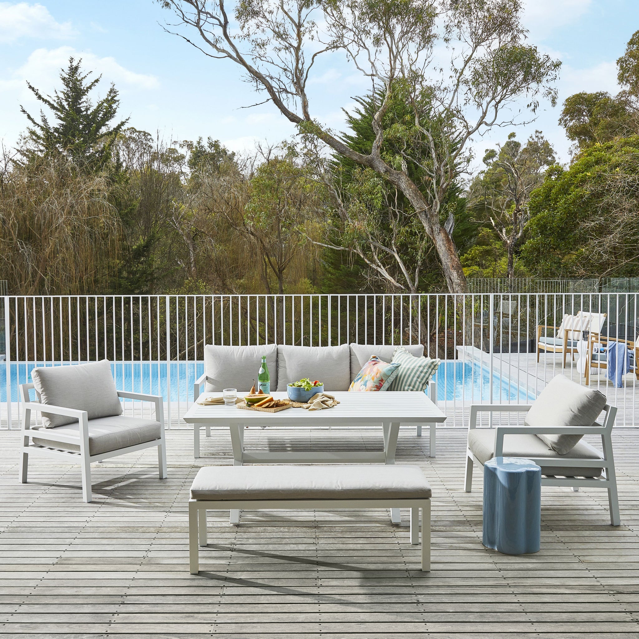 Extended view of the white slatted outdoor dining table and benches set with blue side stool and a pool in background.